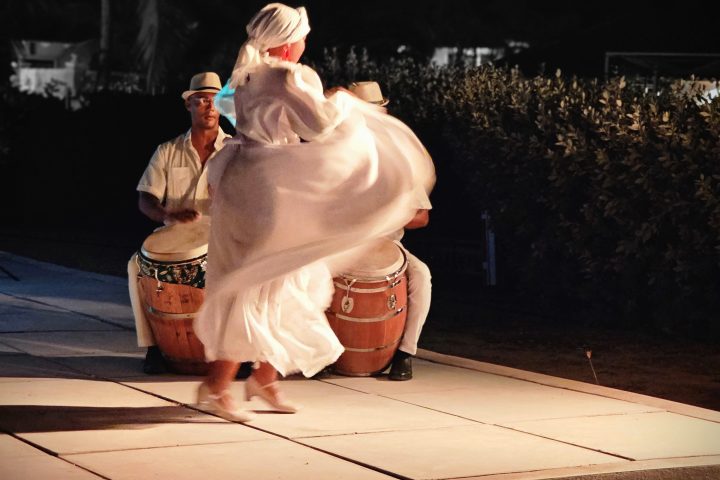 Dancer in white dress twirls on a stage with drummers in the background at night.