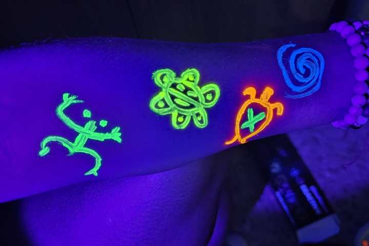 UV-lit arm with neon tribal symbols, including a lizard, flower, turtle, and spiral, adorned with beaded bracelets.